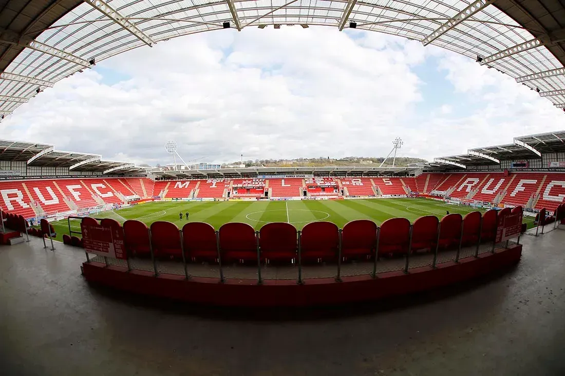 Empty football stadium viewed from the stands, with red seats and a green pitch under a cloudy sky