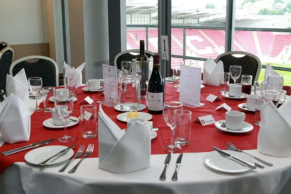 Round banquet table set with red tablecloth, folded napkins, glassware, and bottles by large windows