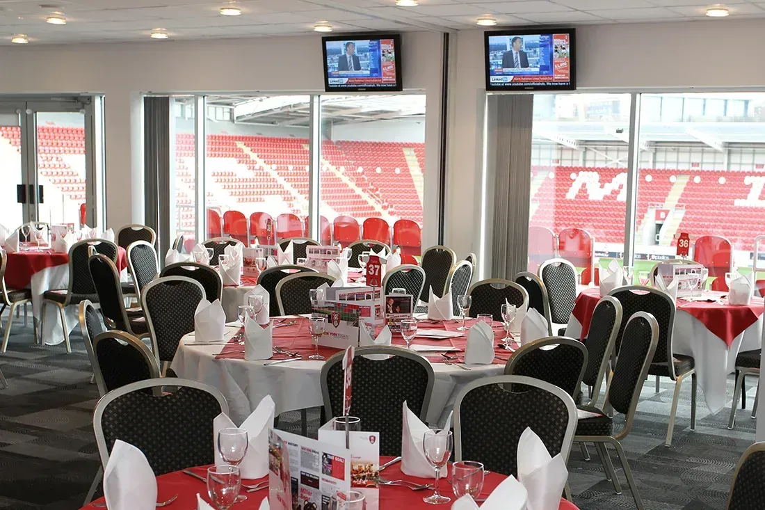 Stadium lounge with round tables, white chairs, and TVs overlooking red seats through large windows
