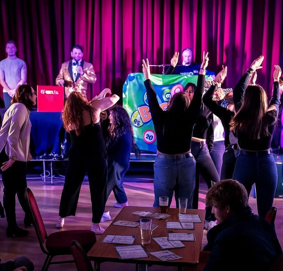 Audience raising hands at a purple-lit stage event with speakers and colorful banners