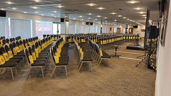 Rows of empty yellow chairs in a large conference hall with stage lighting and equipment.