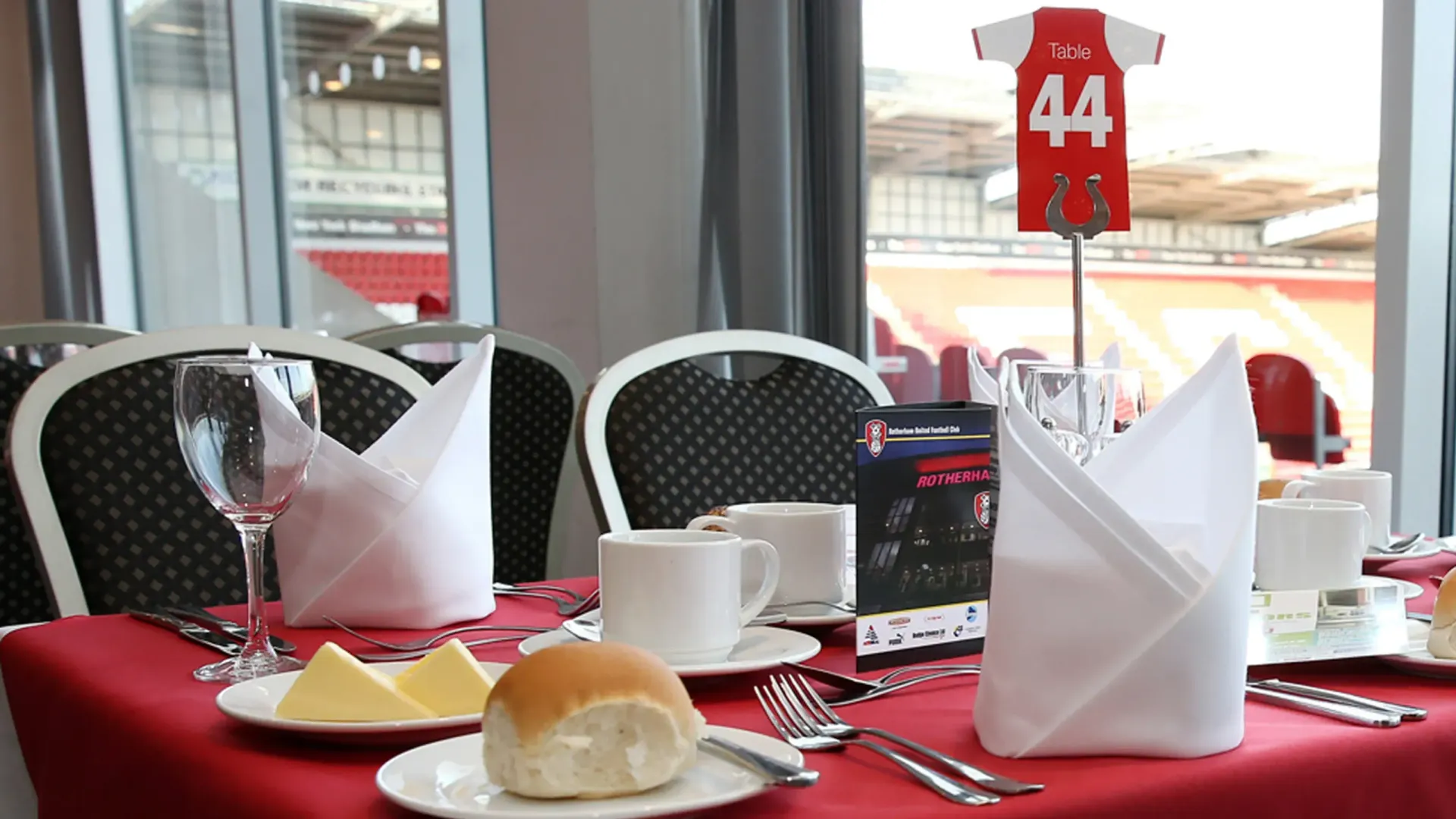 Red table set with cups, napkins, and a bread roll in a windowed café, with a red jersey number 44 in the background