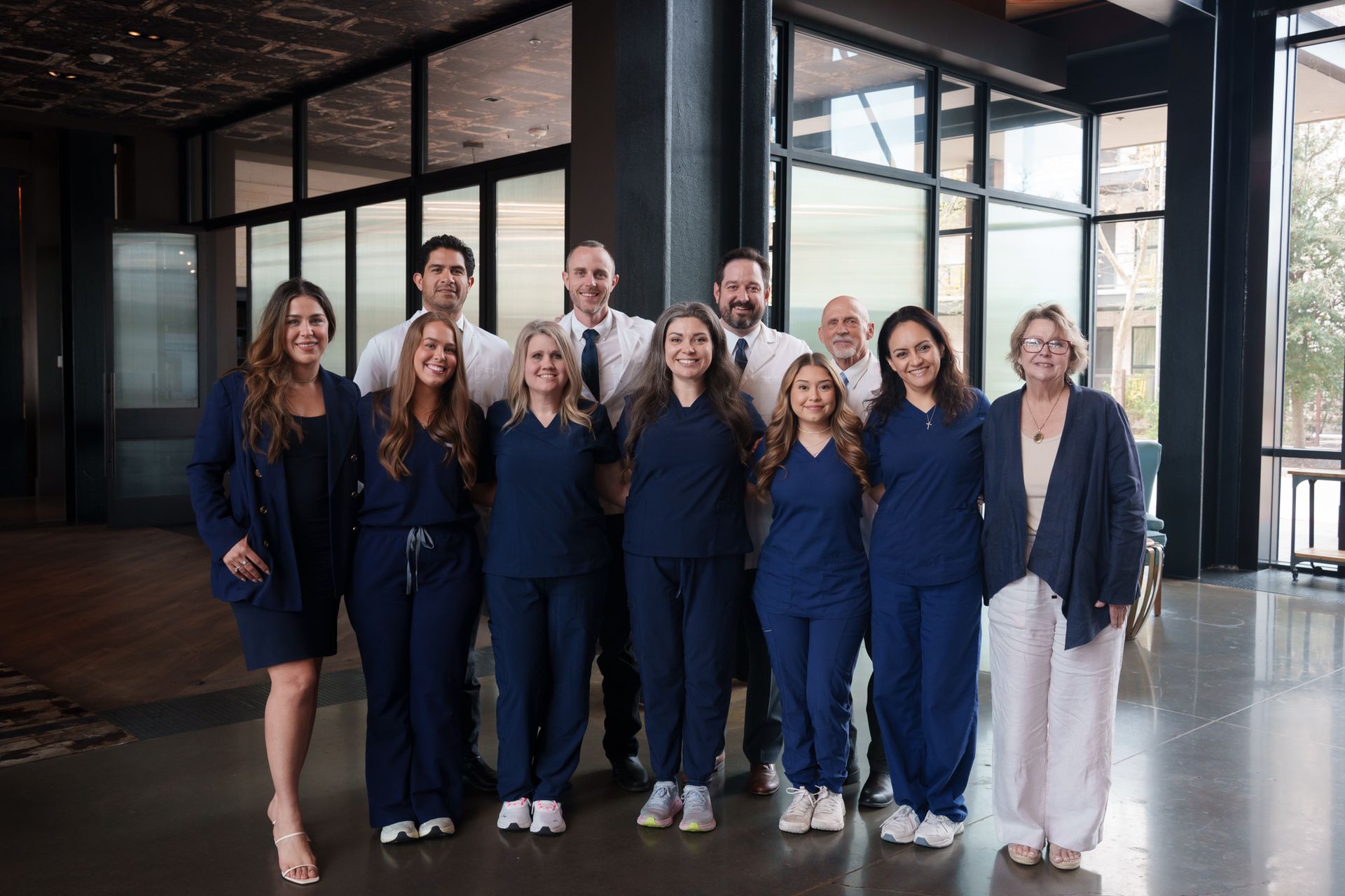 Nine people in navy outfits pose together in a modern glass-walled lobby.