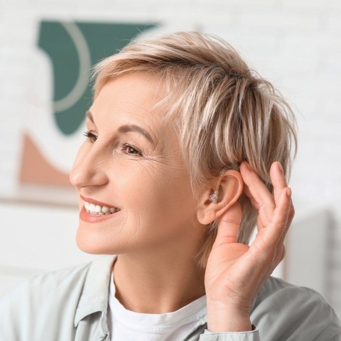 Smiling person with short blonde hair touching an ear in a bright room with abstract wall art