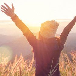 Woman with arms raised, facing a sunset over a field of tall grass.