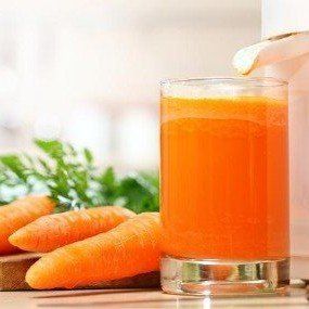 Glass of carrot juice next to carrots and juicer. Bright orange juice, green carrot tops, wooden cutting board.