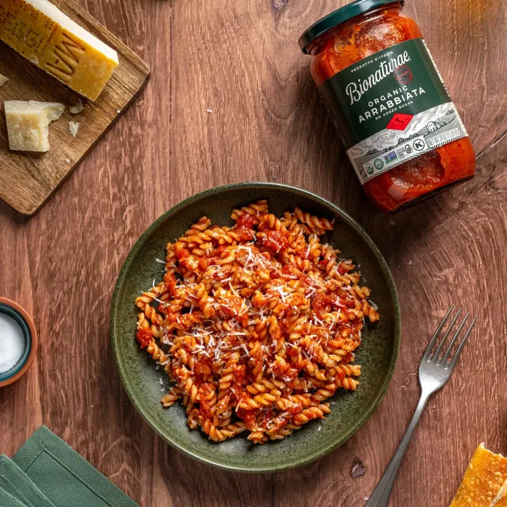 Pasta dish with arrabiata sauce, grated cheese, and jar next to cheese board and fork on wood.