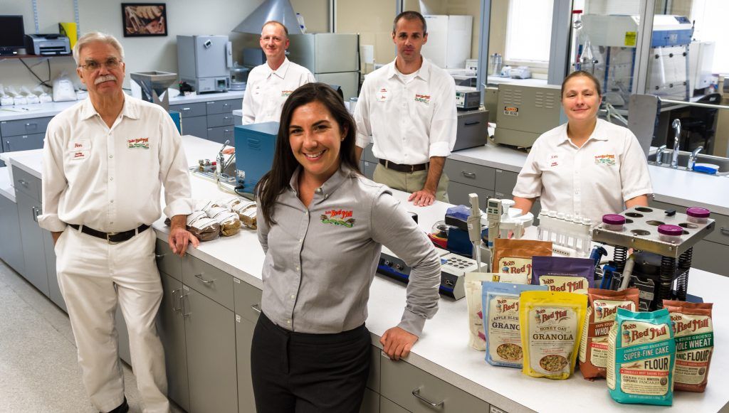 A group of six people in a laboratory setting, some wearing white uniforms and others grey shirts, pose with product packaging on a counter.