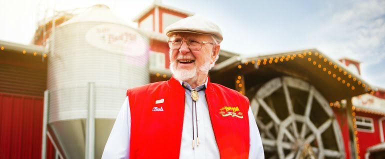 Smiling elderly man in a red vest and cap stands in front of a red building with a water wheel and silo.