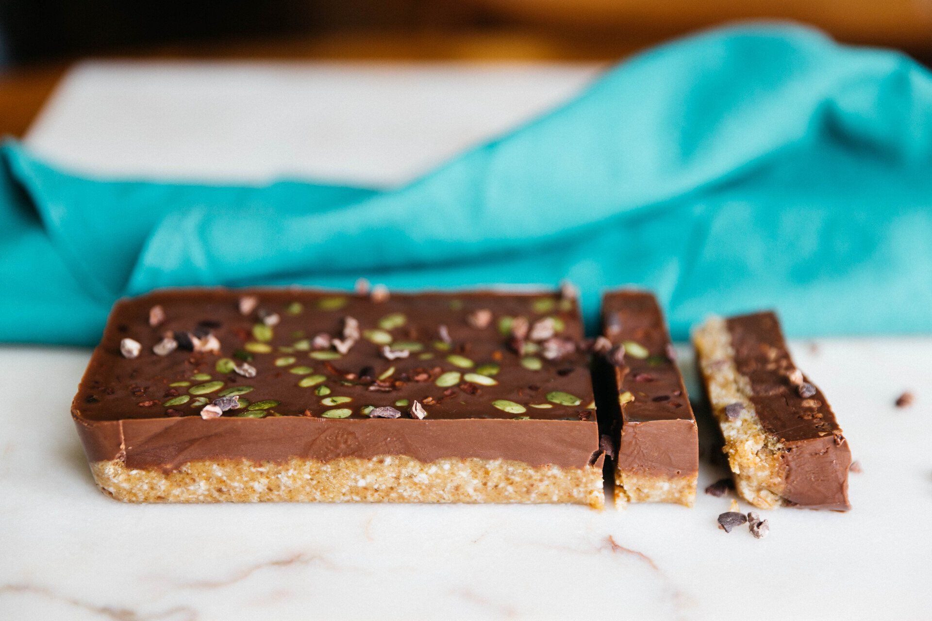 Chocolate dessert bar sliced on a marble surface, topped with cacao nibs and seeds, with a teal cloth in the background.