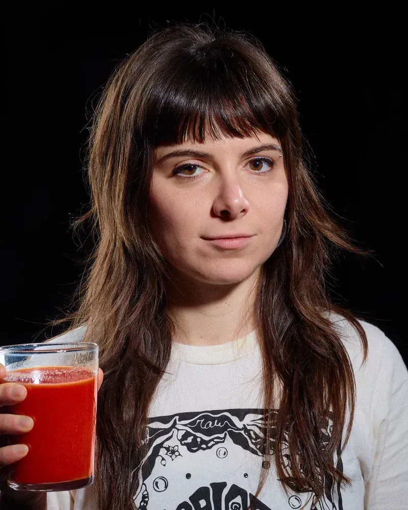 Woman with dark hair and bangs holding a glass of red juice. She wears a white shirt against a black background.