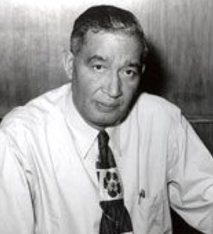 A man with dark hair and a tie, wearing a white shirt, sits in an office setting. He has a neutral expression.