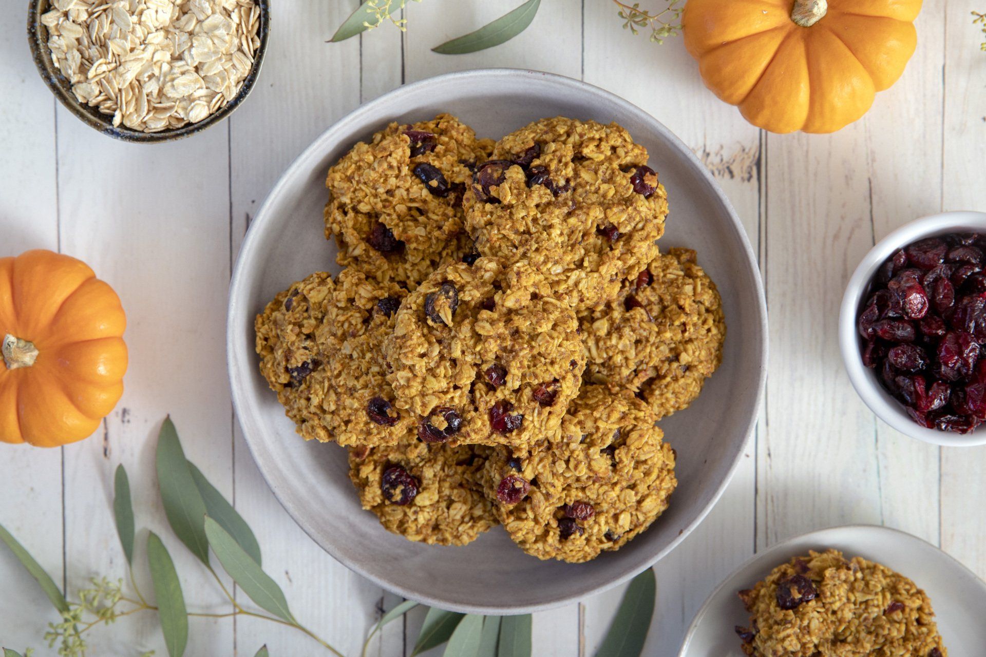 A plate of oatmeal cookies with cranberries, pumpkins, oats, and cranberries arranged on a white wooden surface.