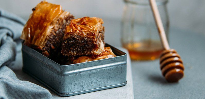 Baklava in a metal tin with honey dipper and jar, on a neutral surface.