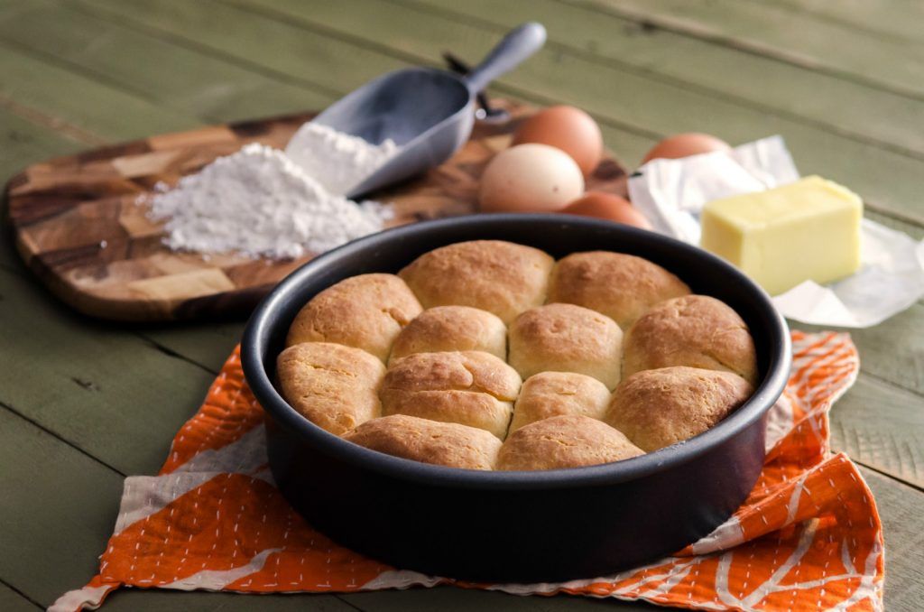 Freshly baked bread rolls in a pan with flour, eggs, and butter on a wooden surface.