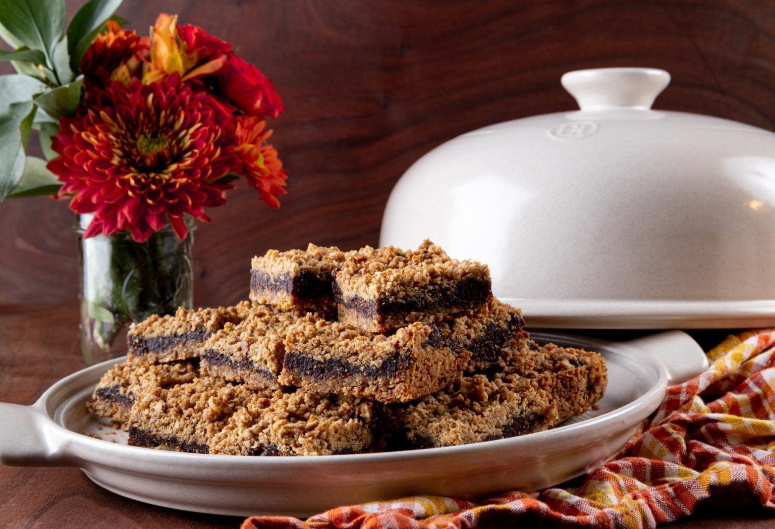 Brownies with crumb topping on a plate, flowers, and a serving dish.