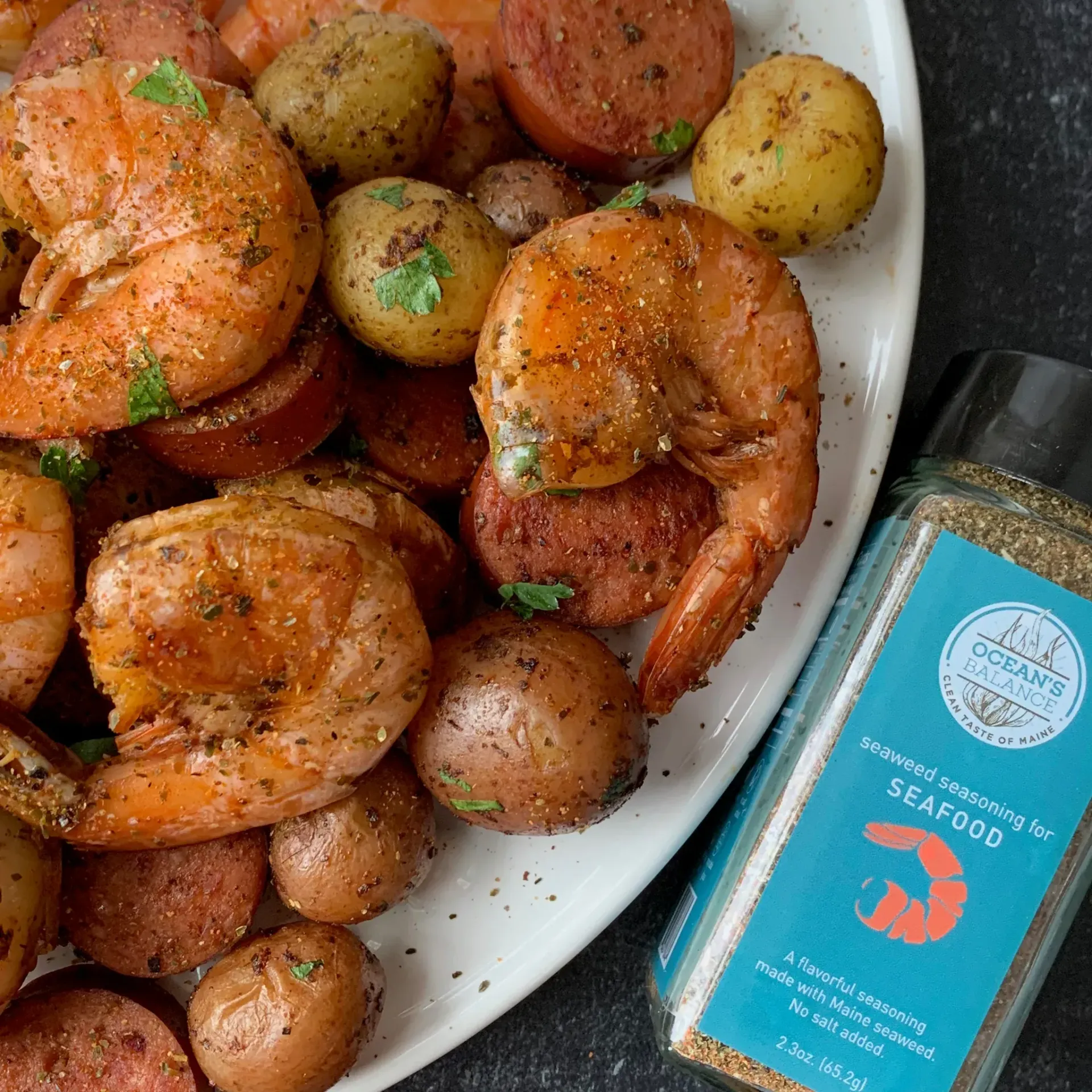 A plate of shrimp boil with shrimp, sausage, and potatoes, seasoned with spices. A seasoning bottle sits nearby.