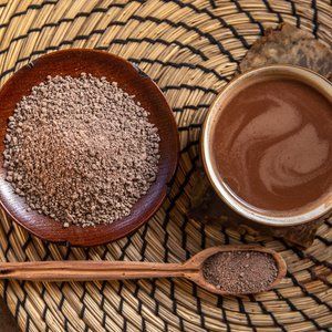 A cup of hot chocolate and a plate of cocoa powder, viewed overhead on a woven placemat. A wooden spoon holds more cocoa powder.