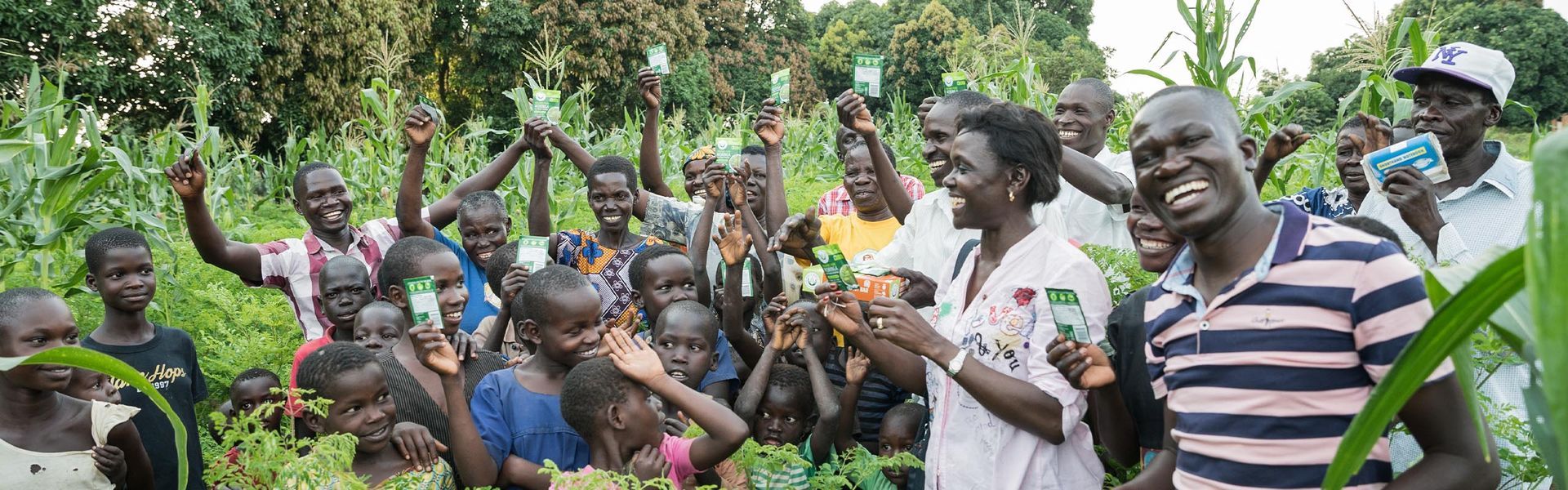 A group of smiling people, mostly dark-skinned, hold up small green objects in a lush field, possibly celebrating a harvest.
