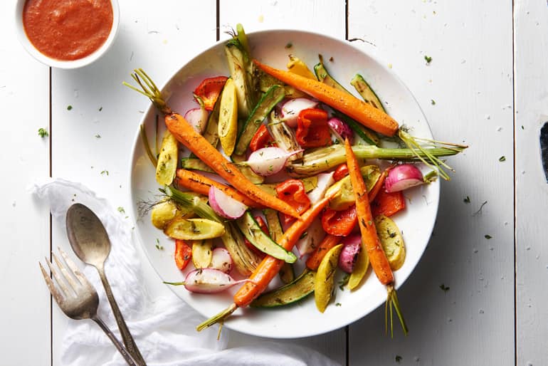 Roasted vegetables, including carrots, radishes, and zucchini, on a white plate with a side of red sauce. White wooden table setting.