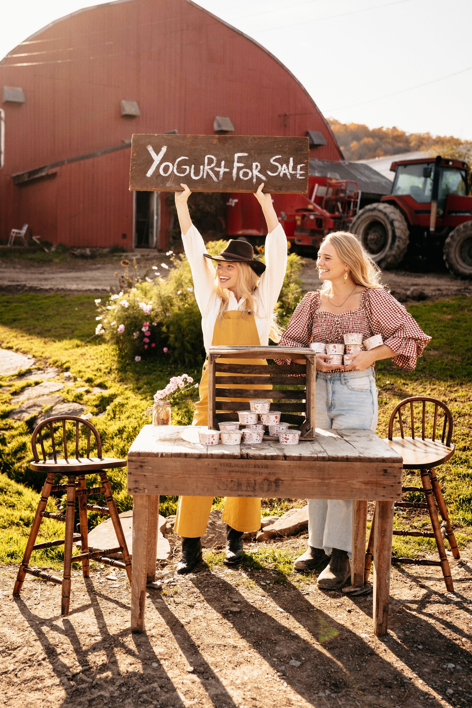 Two young women selling yogurt by a red barn, one holding a 