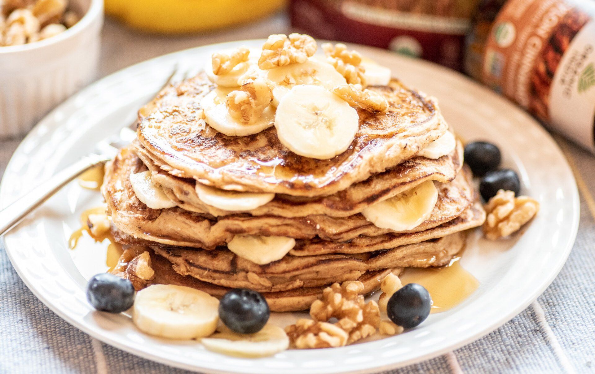 Stack of pancakes topped with banana slices, walnuts, blueberries, and syrup on a white plate.