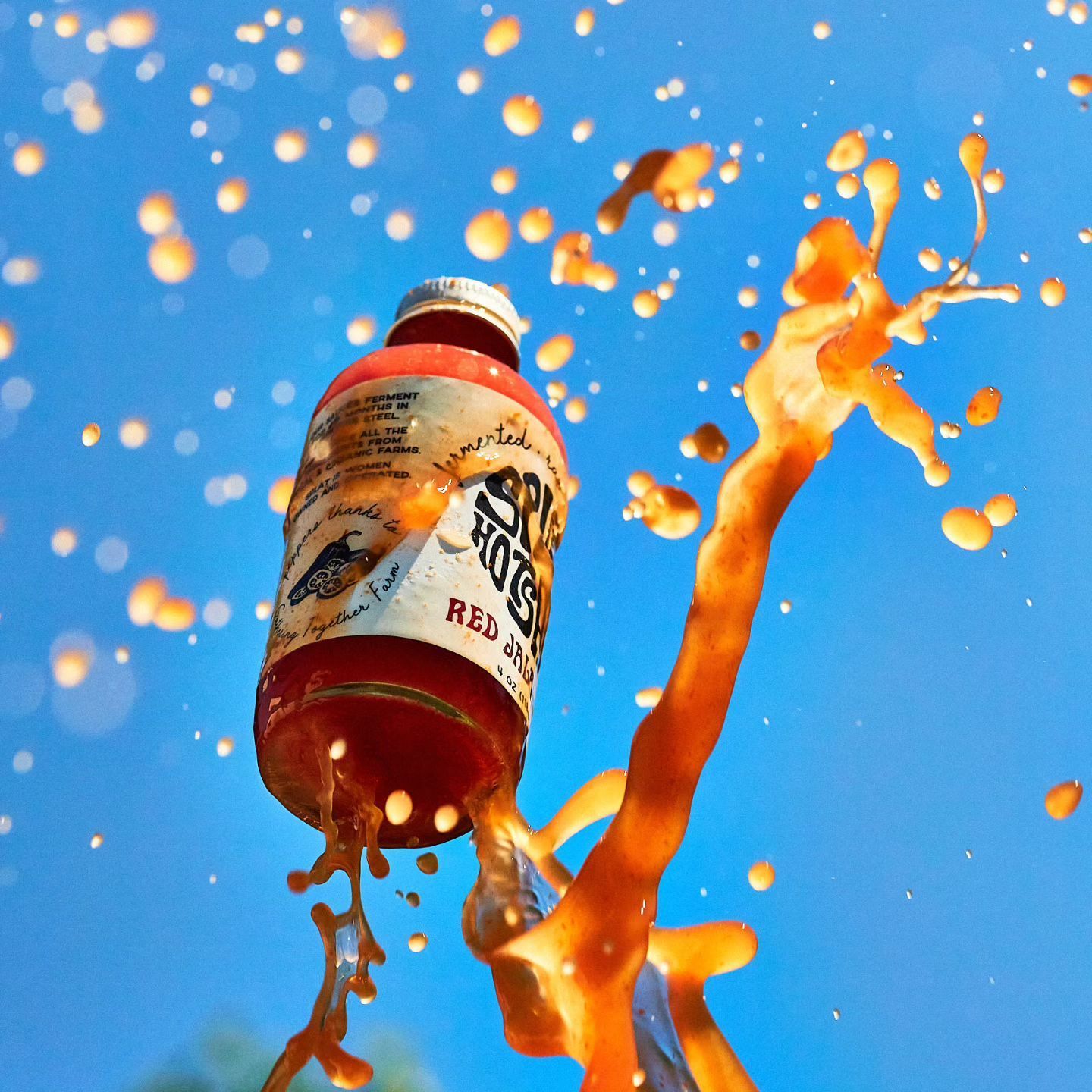 A bottle of hot sauce explodes, spraying red liquid and droplets against a bright blue sky.