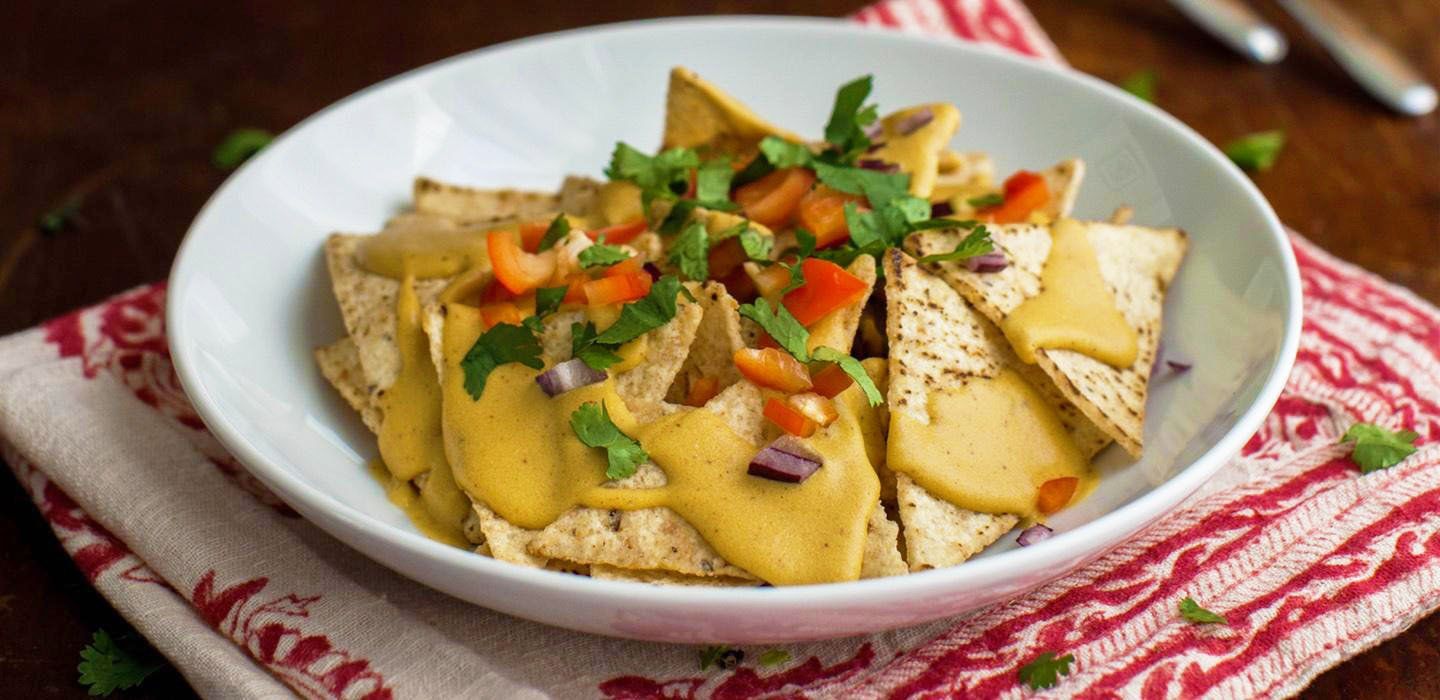 Nachos in a white bowl, topped with orange sauce, chopped red peppers, and green herbs. The dish sits on a red and white striped cloth.