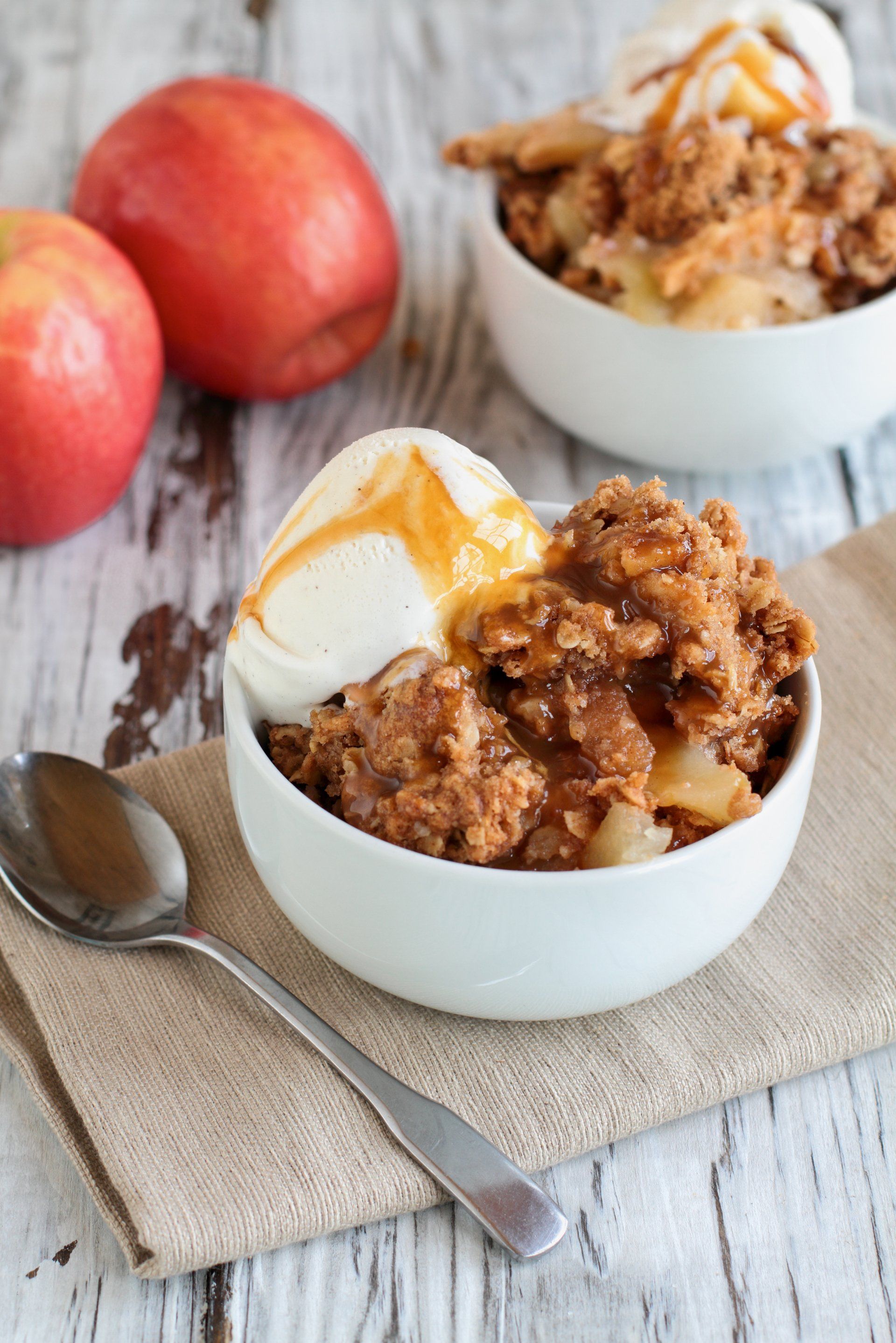 Apple crisp with ice cream and caramel sauce, served in a white bowl, apples in background.