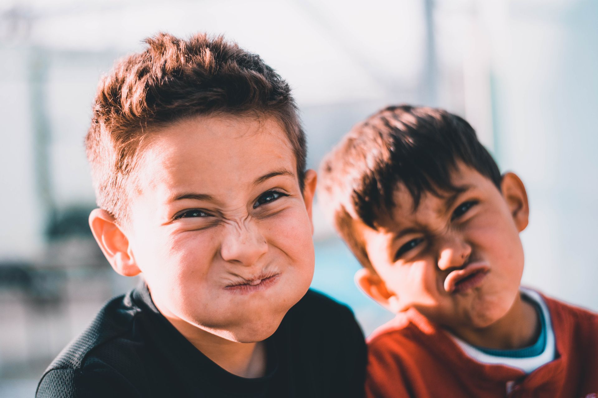 Two young boys making silly faces, both with squinched eyes and puckered lips, outdoors.