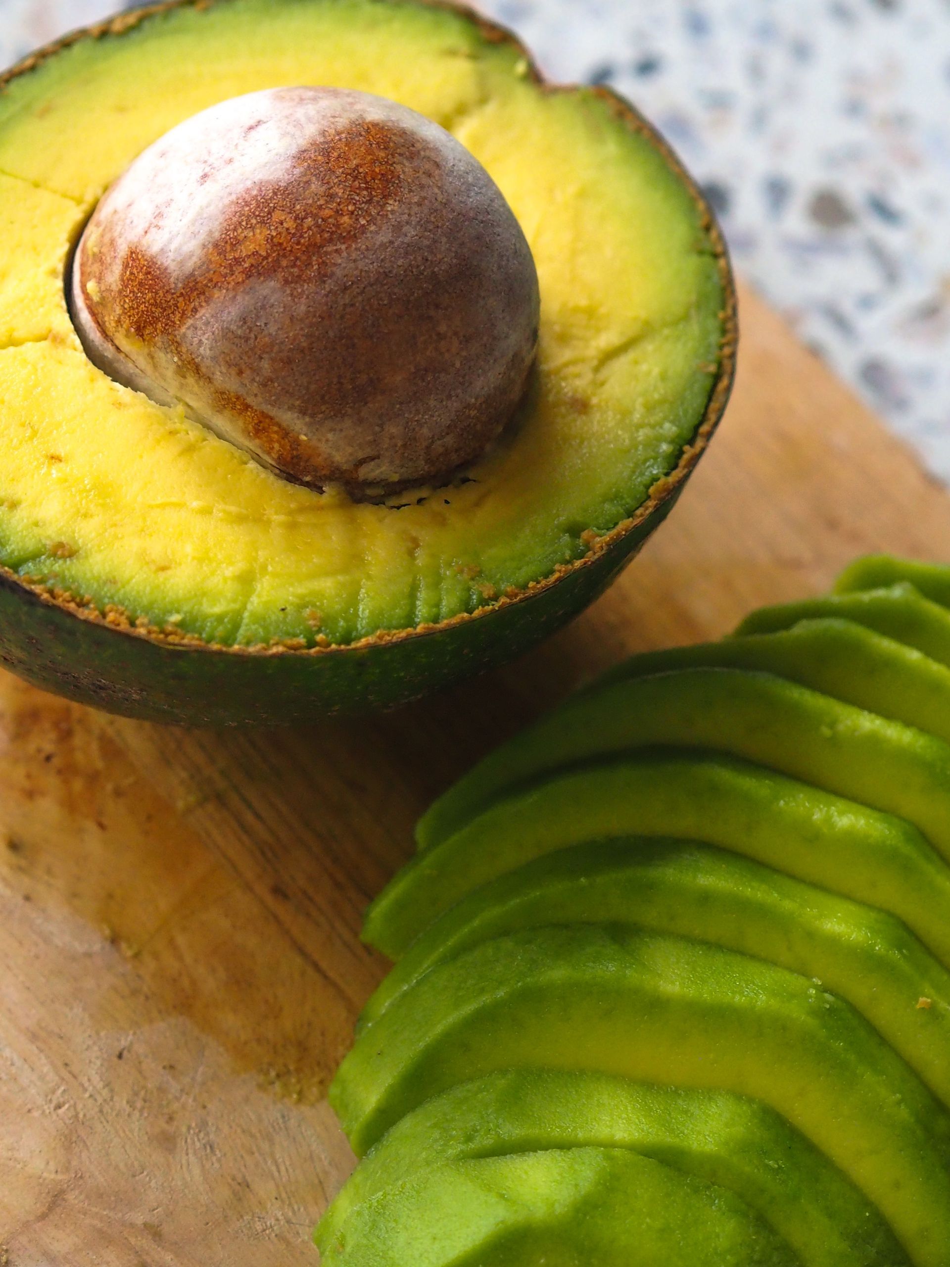 Sliced avocado on a wooden cutting board, one half with the pit, other half sliced.