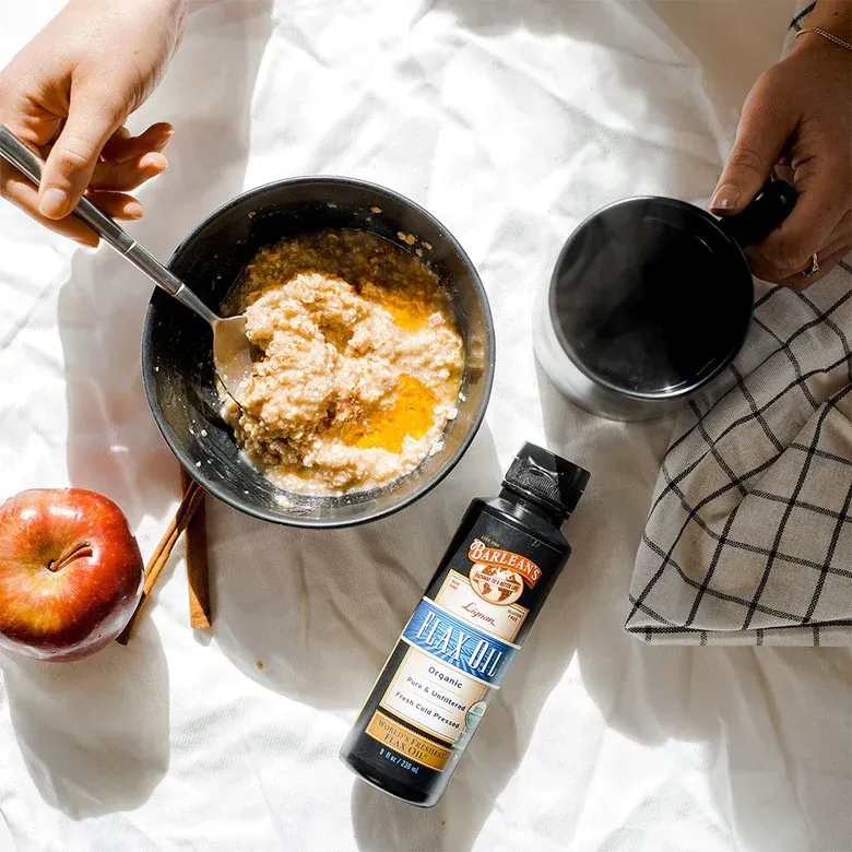 Overhead view: Oatmeal in a bowl with flax oil, coffee, apple, and hands on a bed.