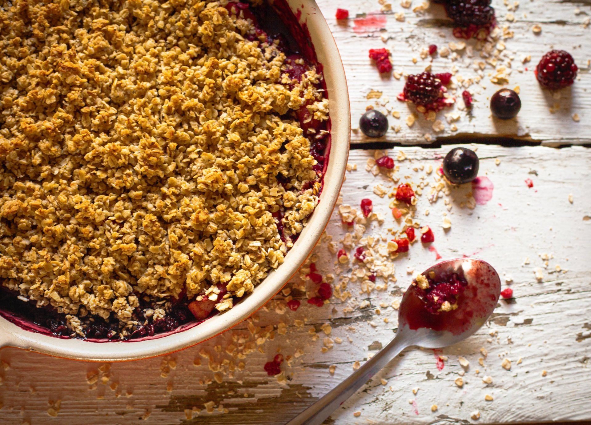 Berry crumble in a round dish, with a spoonful of the filling, scattered berries, and oats on a rustic white wooden surface.