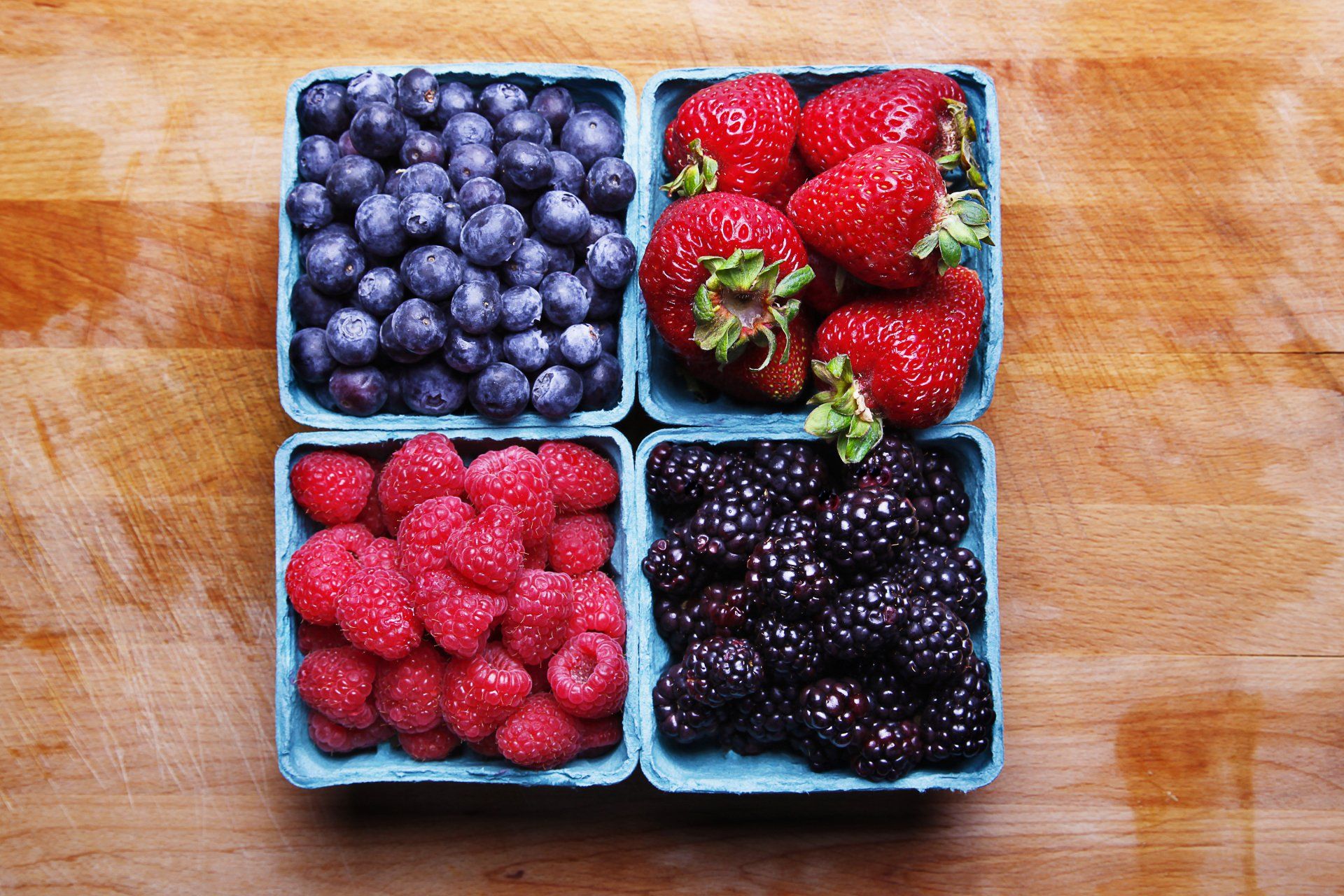 Four containers of fresh berries: blueberries, strawberries, raspberries, and blackberries on a wooden surface.