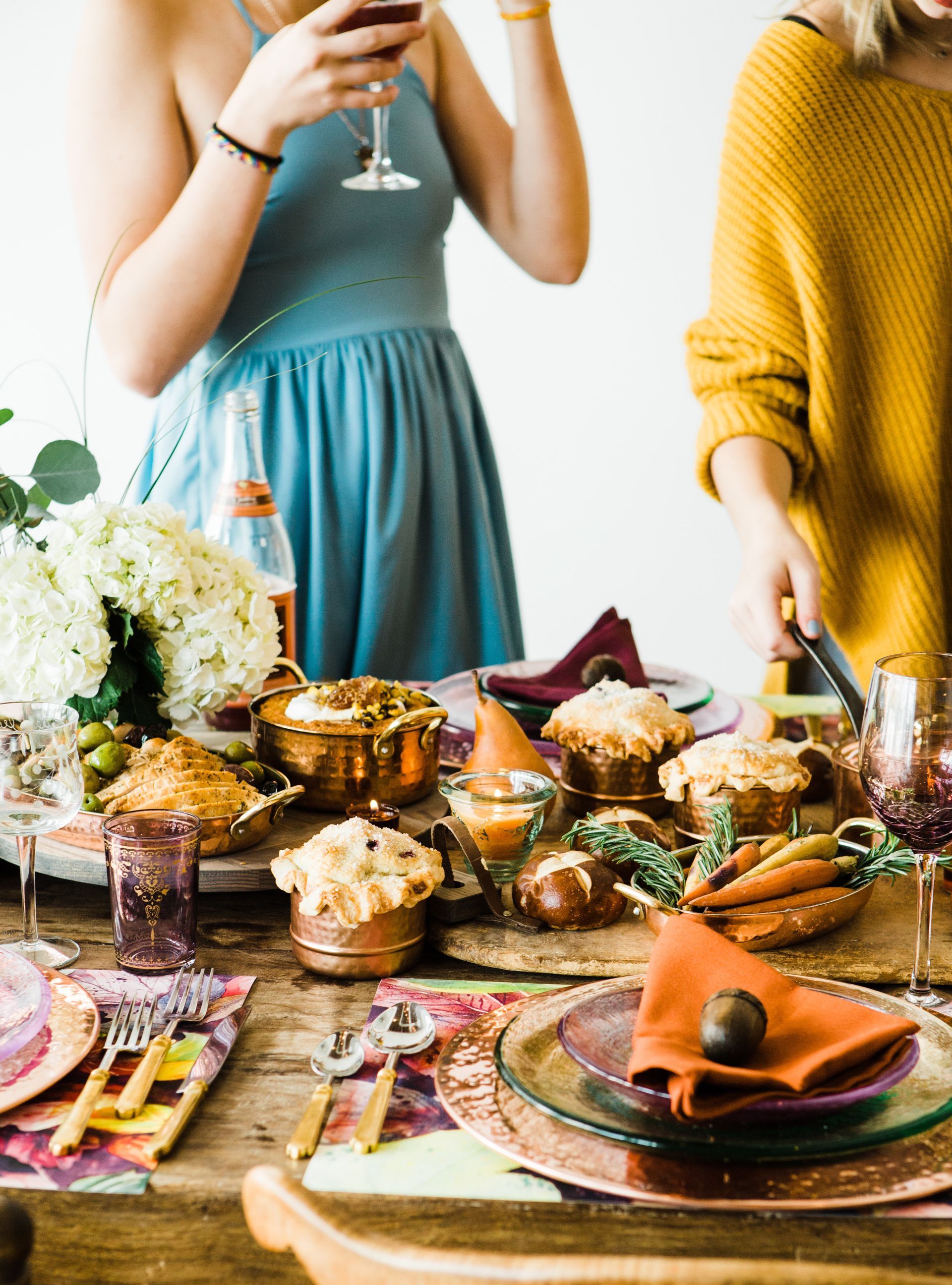 Friends at a festive table, food spread. Woman in blue drinks, other in yellow serves; food is autumnal tones.
