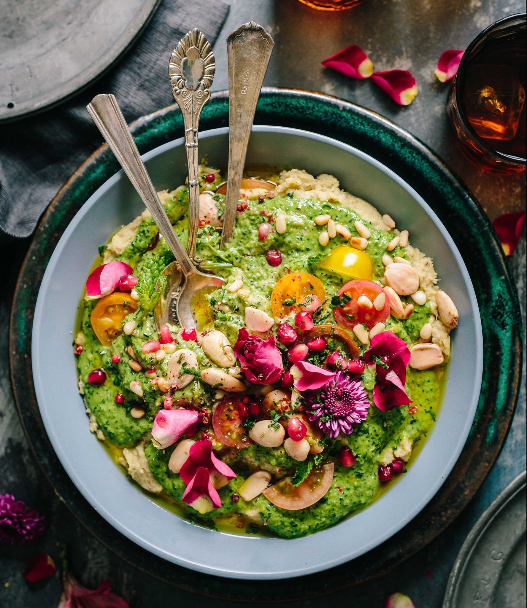 Close-up of a vibrant, garnished dish in a blue bowl. The dish features a green sauce topped with beans, nuts, tomatoes, flowers, and pomegranate seeds.