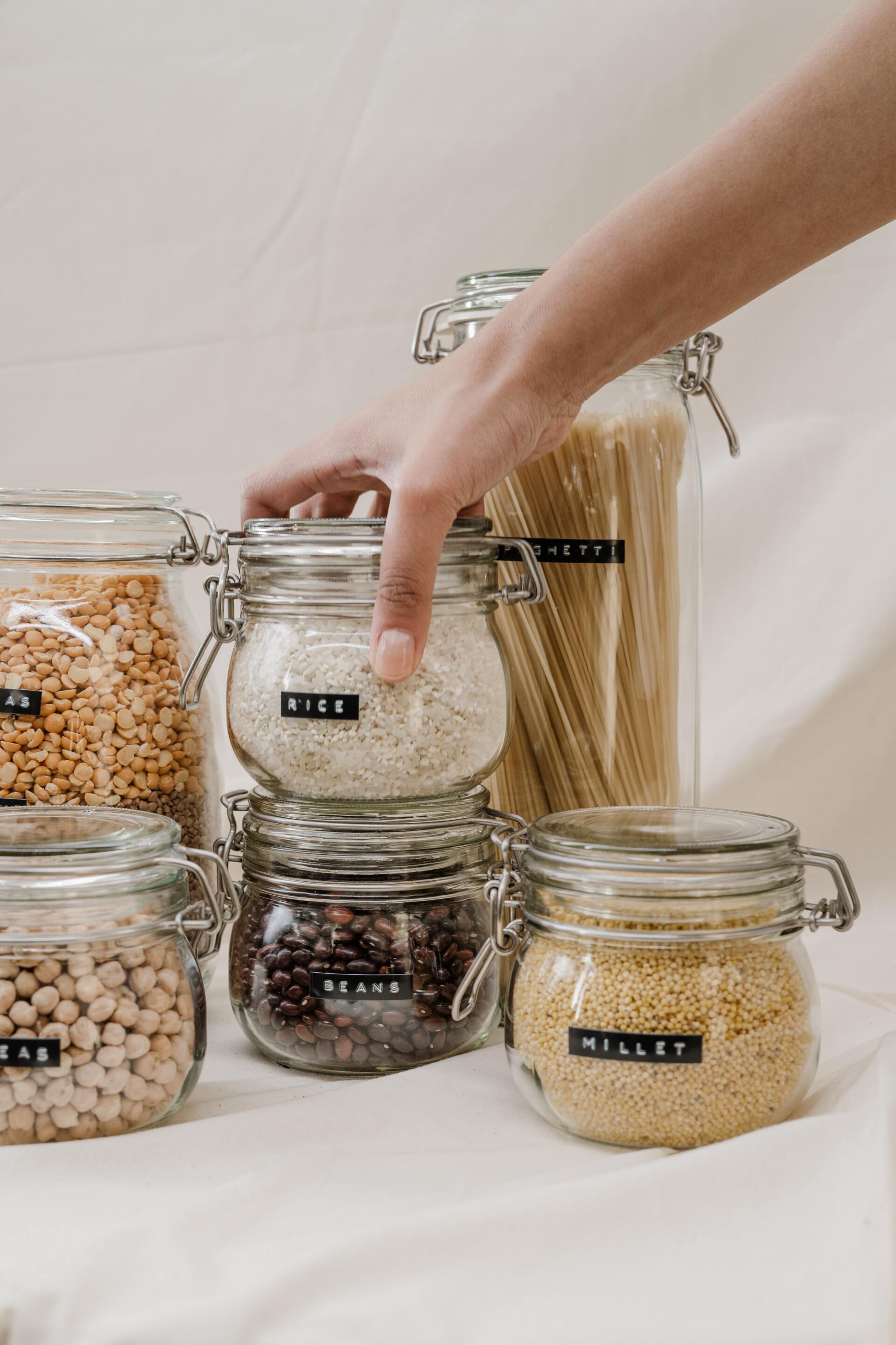Person's hand reaching for a jar of rice. The jars hold various dry goods, each labeled, arranged on a white surface.