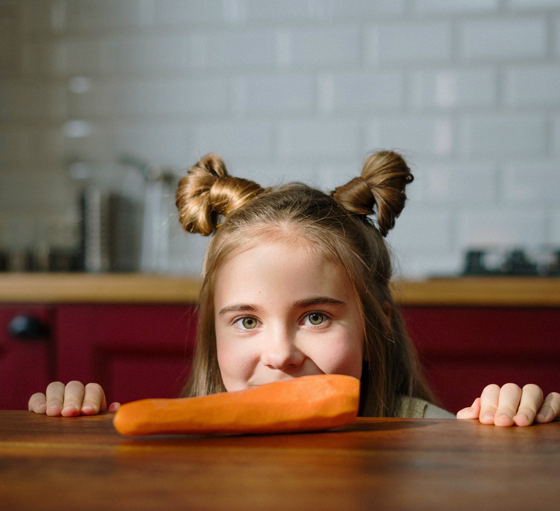 Girl with space buns smiles, holding a carrot on a wooden table in a kitchen.