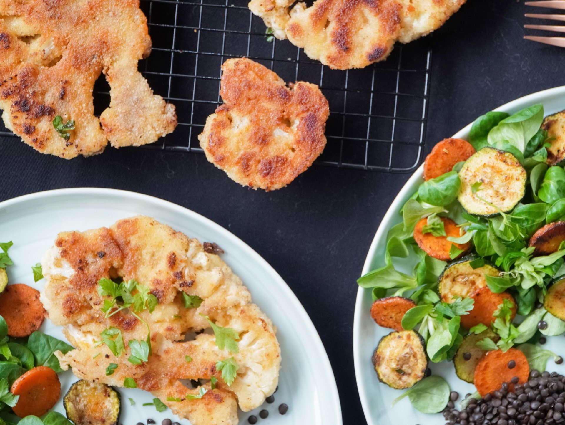 Breaded cauliflower steaks with roasted vegetables and salad, arranged on plates and a cooling rack.