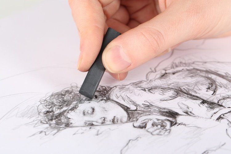 Hand using a charcoal stick to smudge a charcoal sketch of a person's face and upper body on white paper.