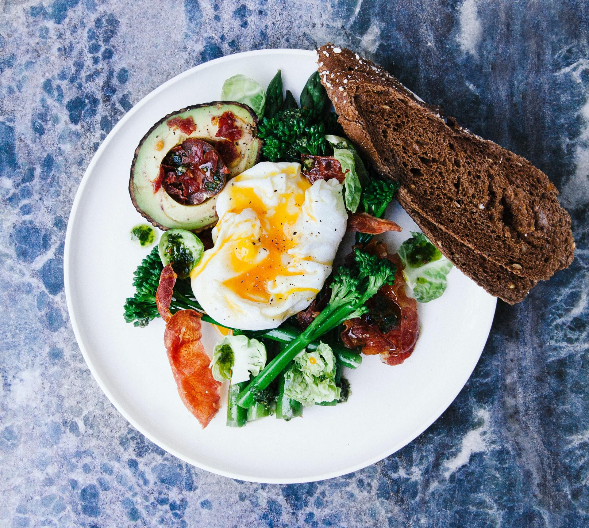 Plate of brunch: poached egg with runny yolk, bacon, avocado with salsa, greens, and rye bread.