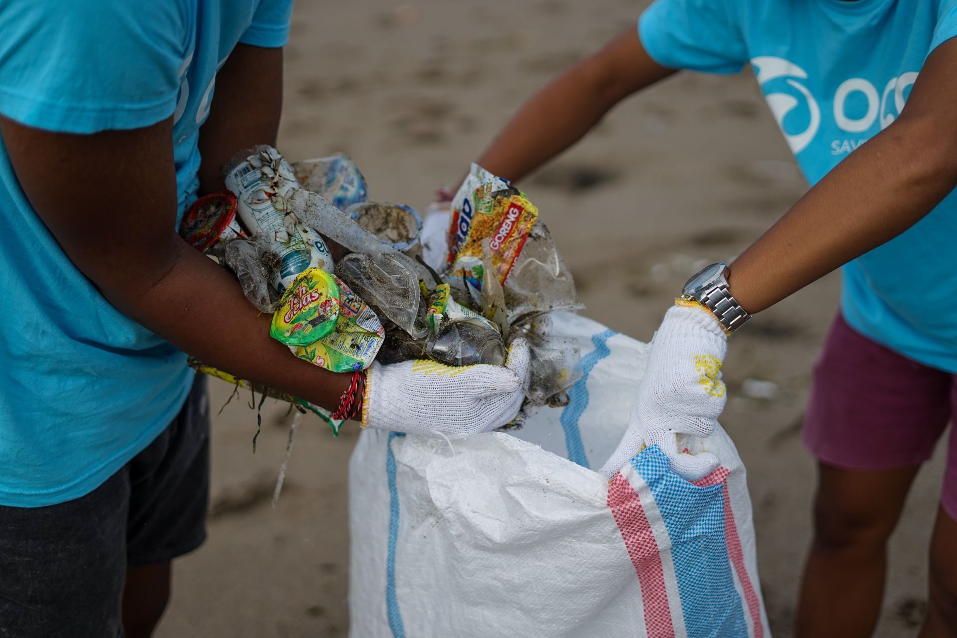 Two people in blue shirts collecting trash on a beach, holding a white bag full of plastic waste.
