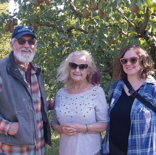Three people smiling in front of a fruit tree. A man in a blue cap and vest, a woman with blonde hair, and a younger woman in a blue plaid shirt.