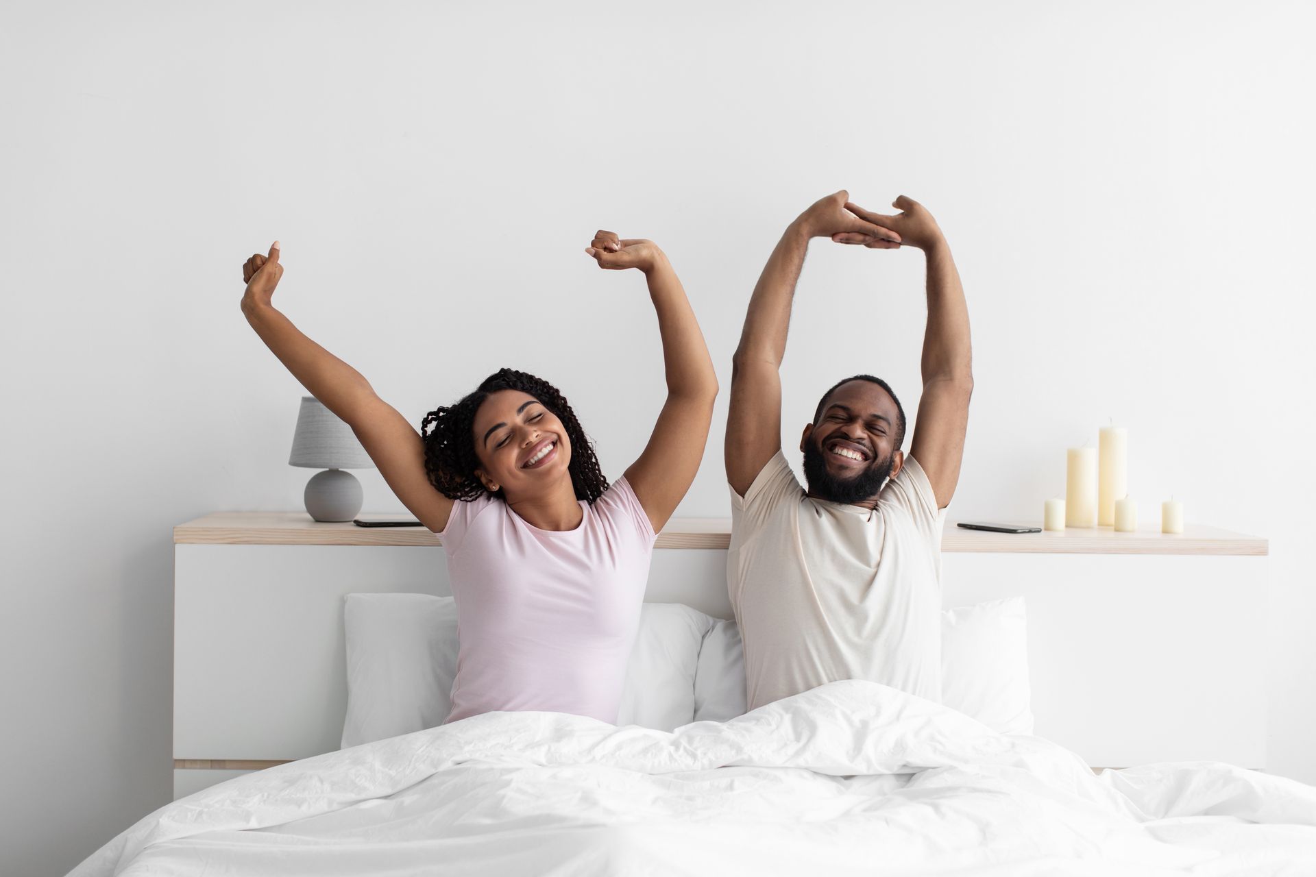 Smiling couple stretching in bed, arms raised. They are in a bright bedroom with white walls and bedding.