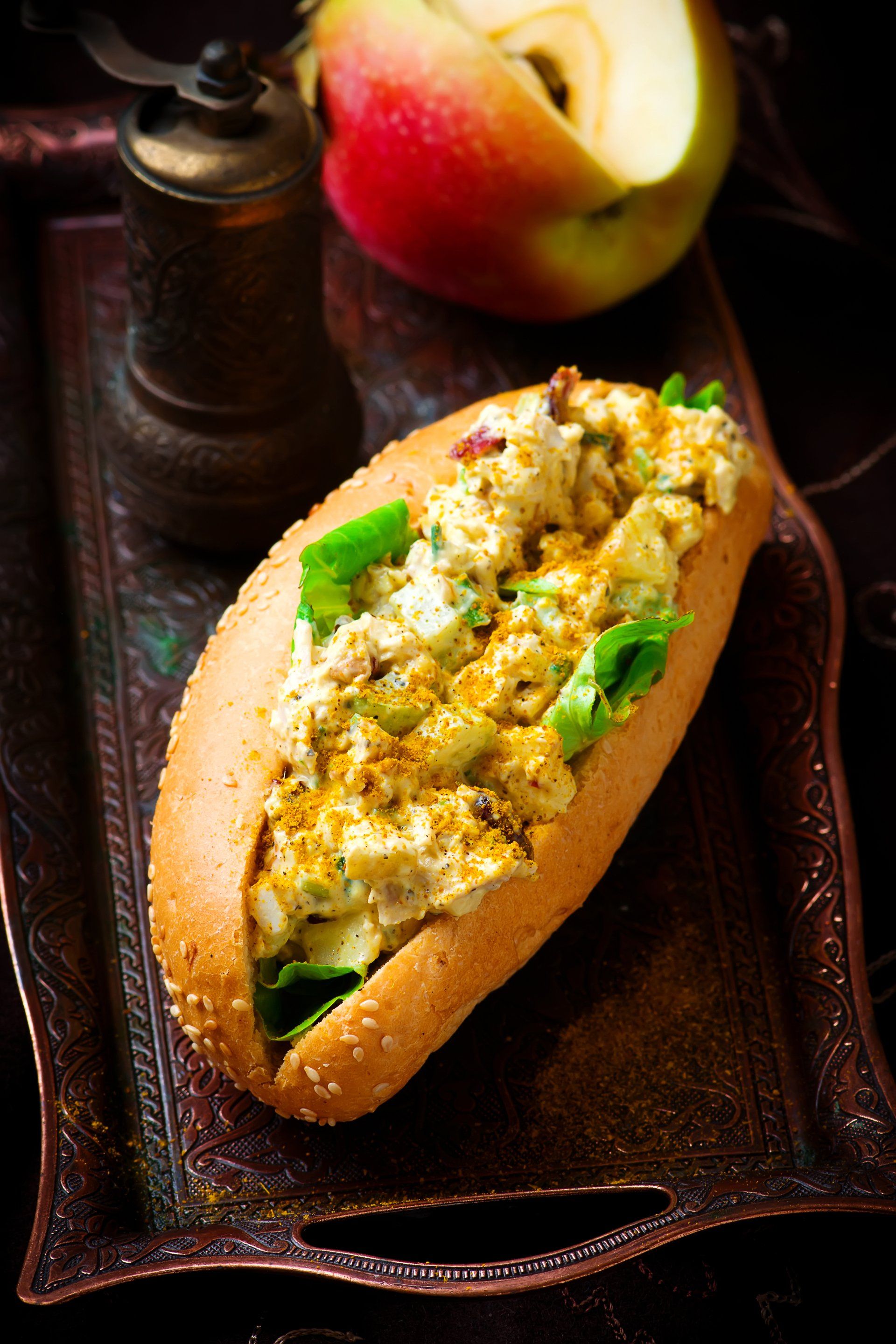 Chicken salad sandwich on a sesame bun, served on a decorative tray with an apple and pepper grinder.