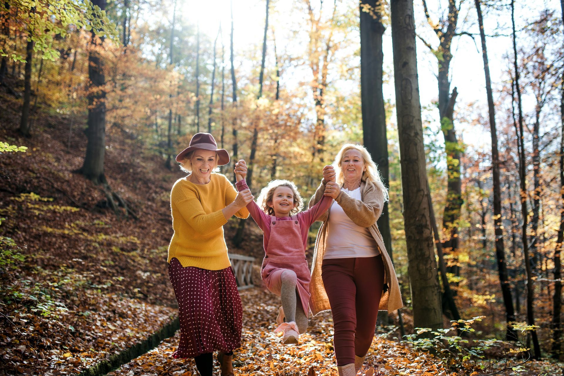 Three generations in a sunny forest: a girl swings between a smiling grandmother and great-grandmother. Autumn leaves and trees surround them.