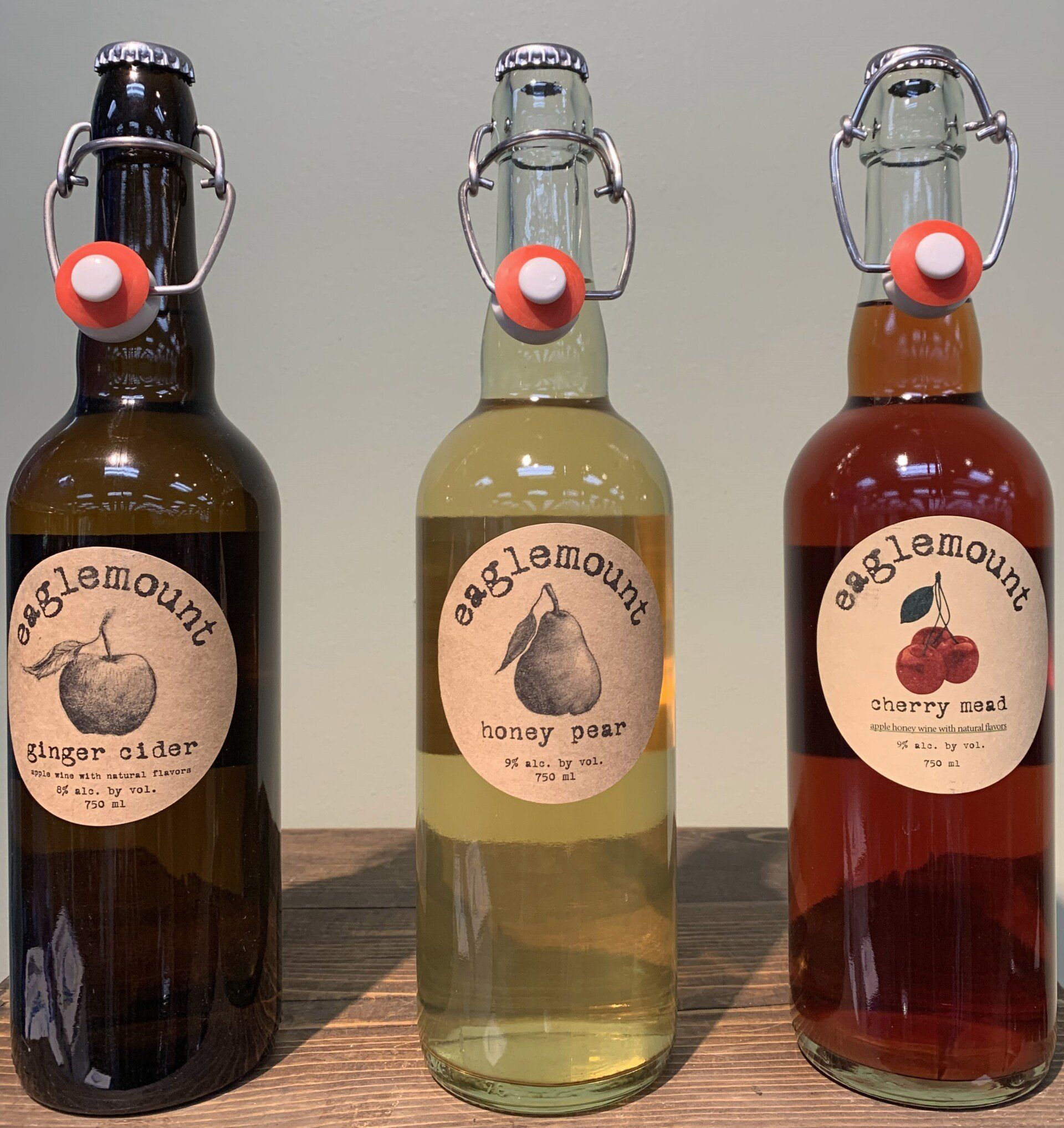 Three glass bottles of Eaglemount artisan drinks: ginger cider, honey pear, and cherry swirl. Each has a swing-top closure and a round label.
