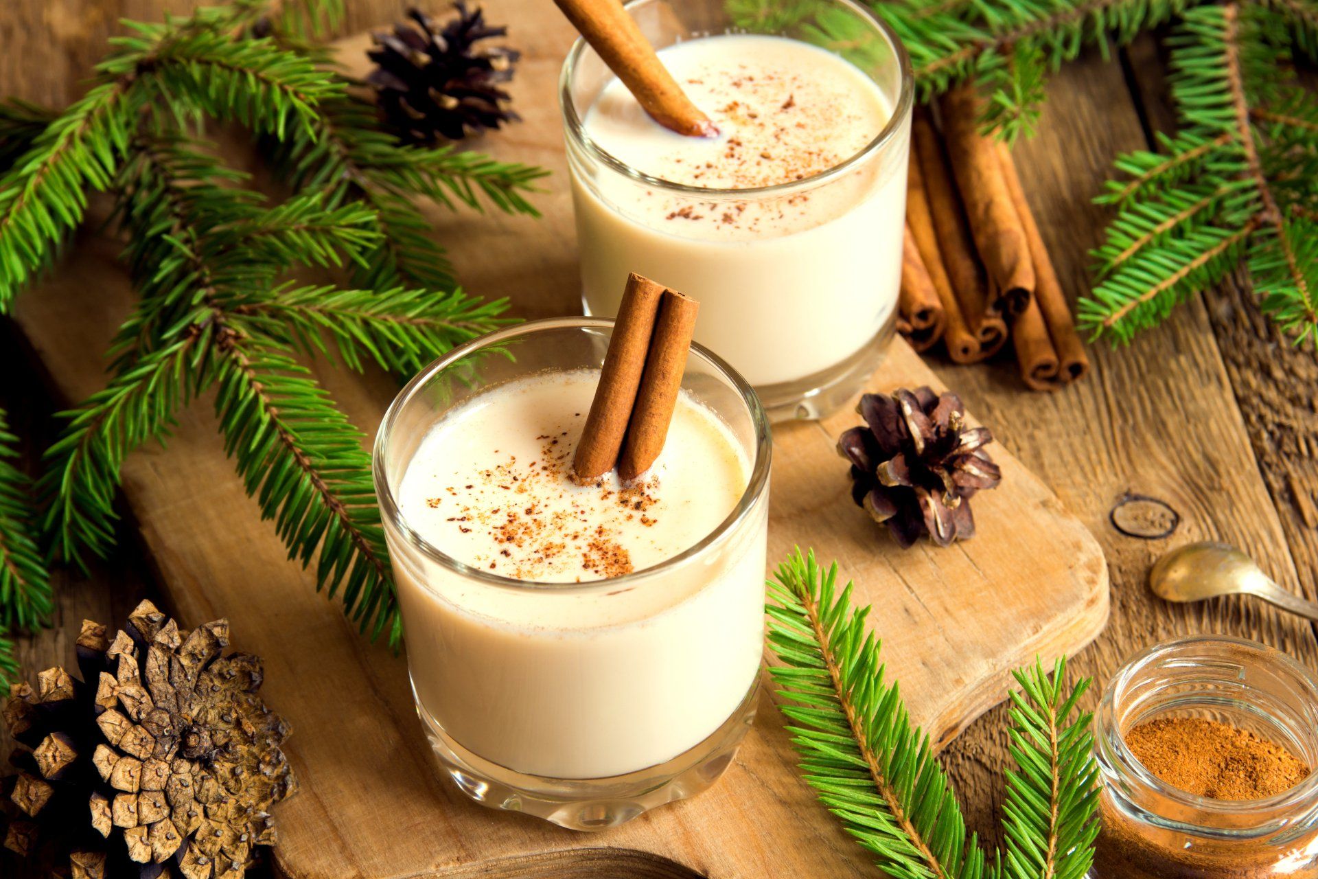 Two glasses of eggnog, garnished with cinnamon sticks and spice, sit on a wooden board with pine boughs and pinecones.