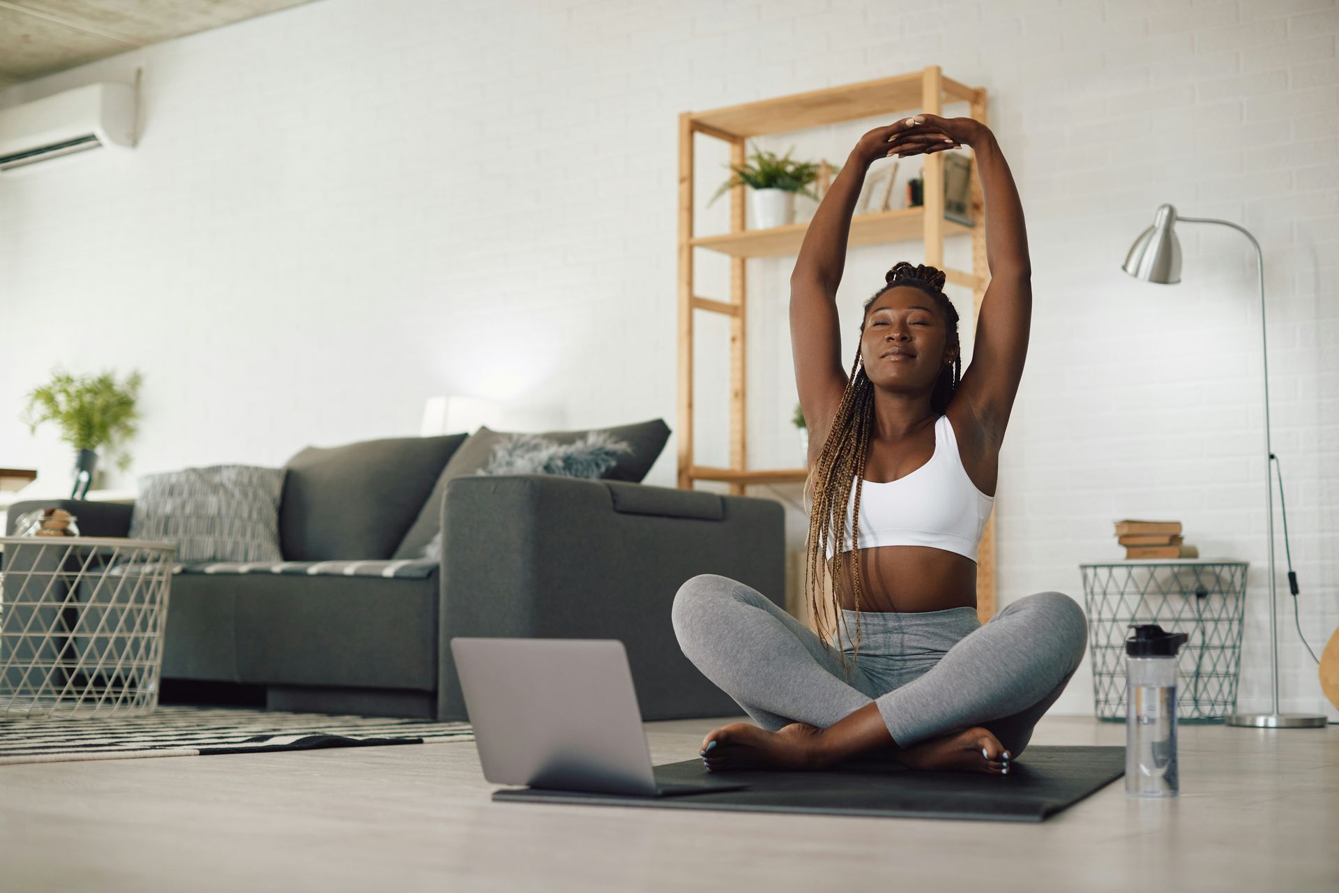 Woman in workout clothes stretches arms overhead while meditating in front of a laptop at home.