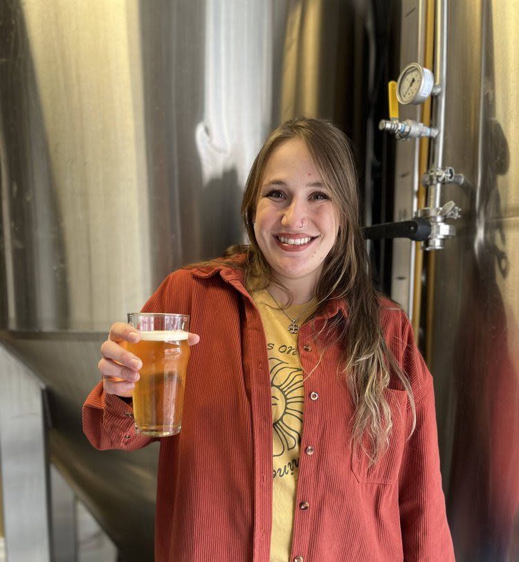 Woman in a reddish-orange shirt smiling and holding a glass of beer in front of a brewery tank.